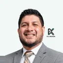 Retrato de John Cevallos, sonriente, con traje gris y corbata de rayas, sobre fondo claro con el logo de Klinoda