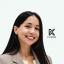 Retrato de Norka Monge, sonriente, con chaqueta beige y blusa blanca, sobre fondo claro con el logo de Klinoda
